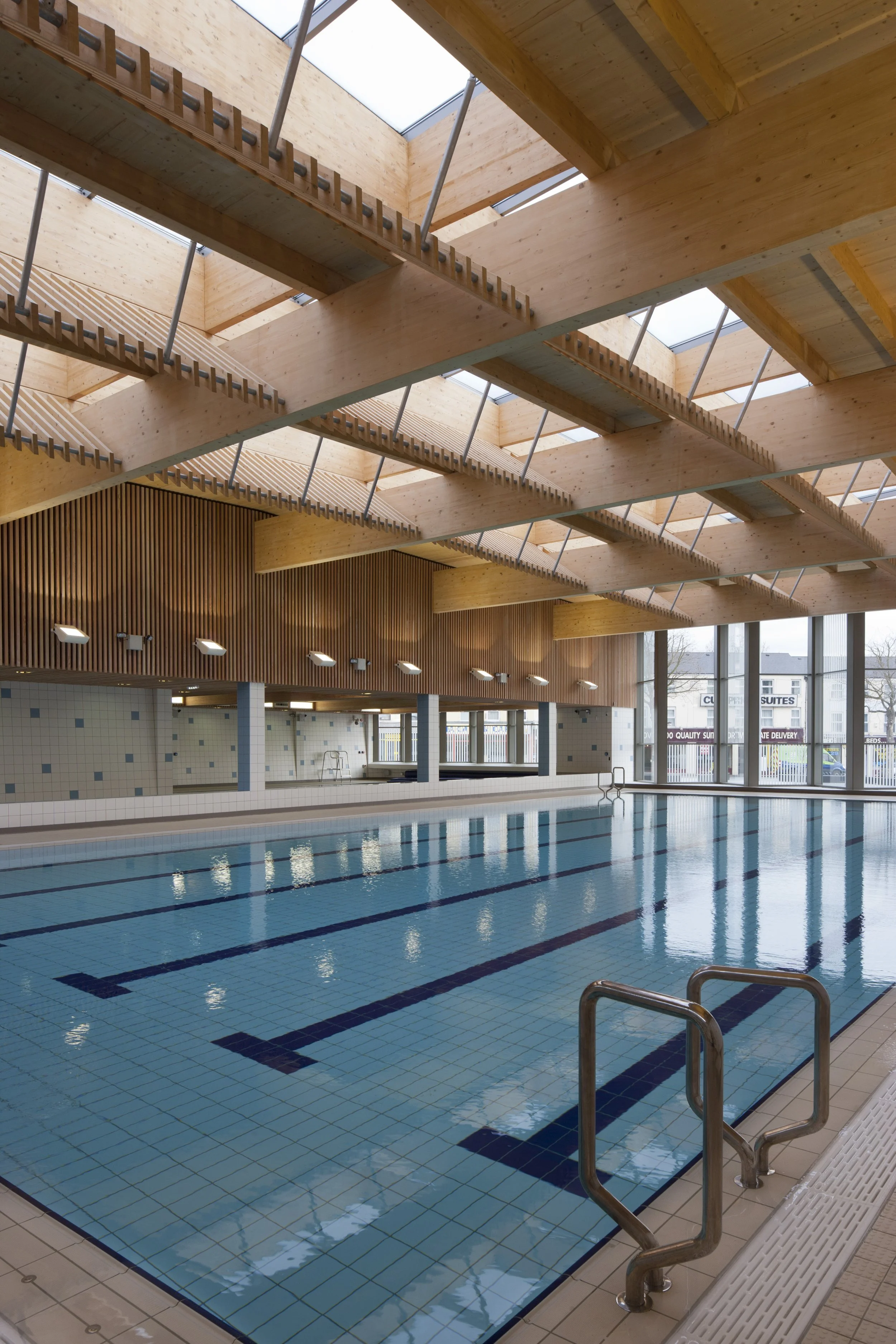 Levitate Architects' Victoria Leisure Centre: A wide architectural shot of the empty swimming pool lanes showing the extensive glulam timber roof structure and large floor-to-ceiling windows.