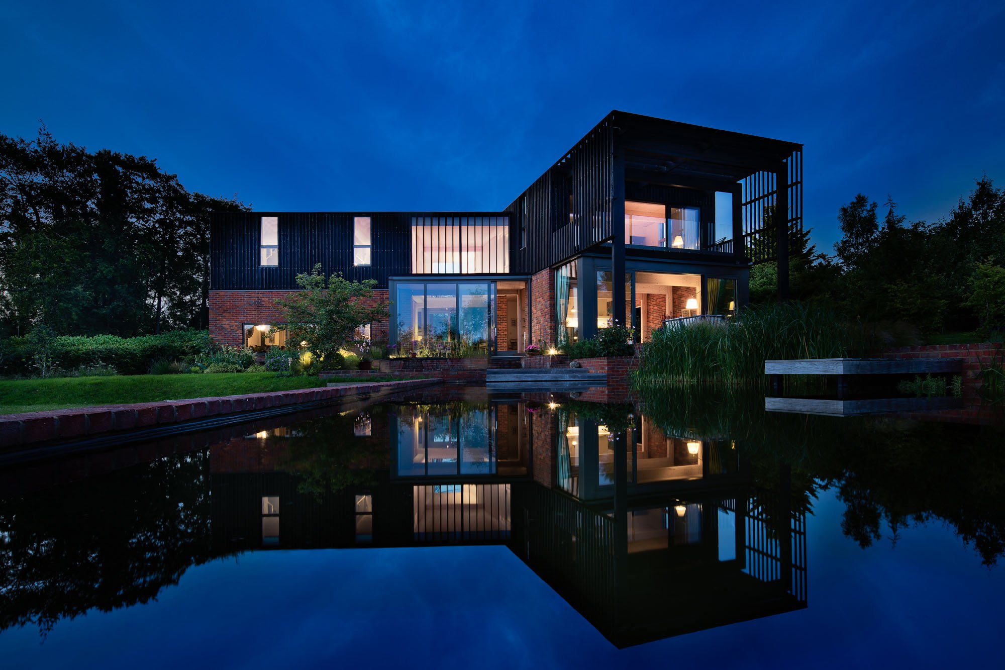 Dusk view of the exterior of Levitate's mid-century modern inspired two-story house with black timber and brick cladding, with a natural swimming lake in the foreground.
