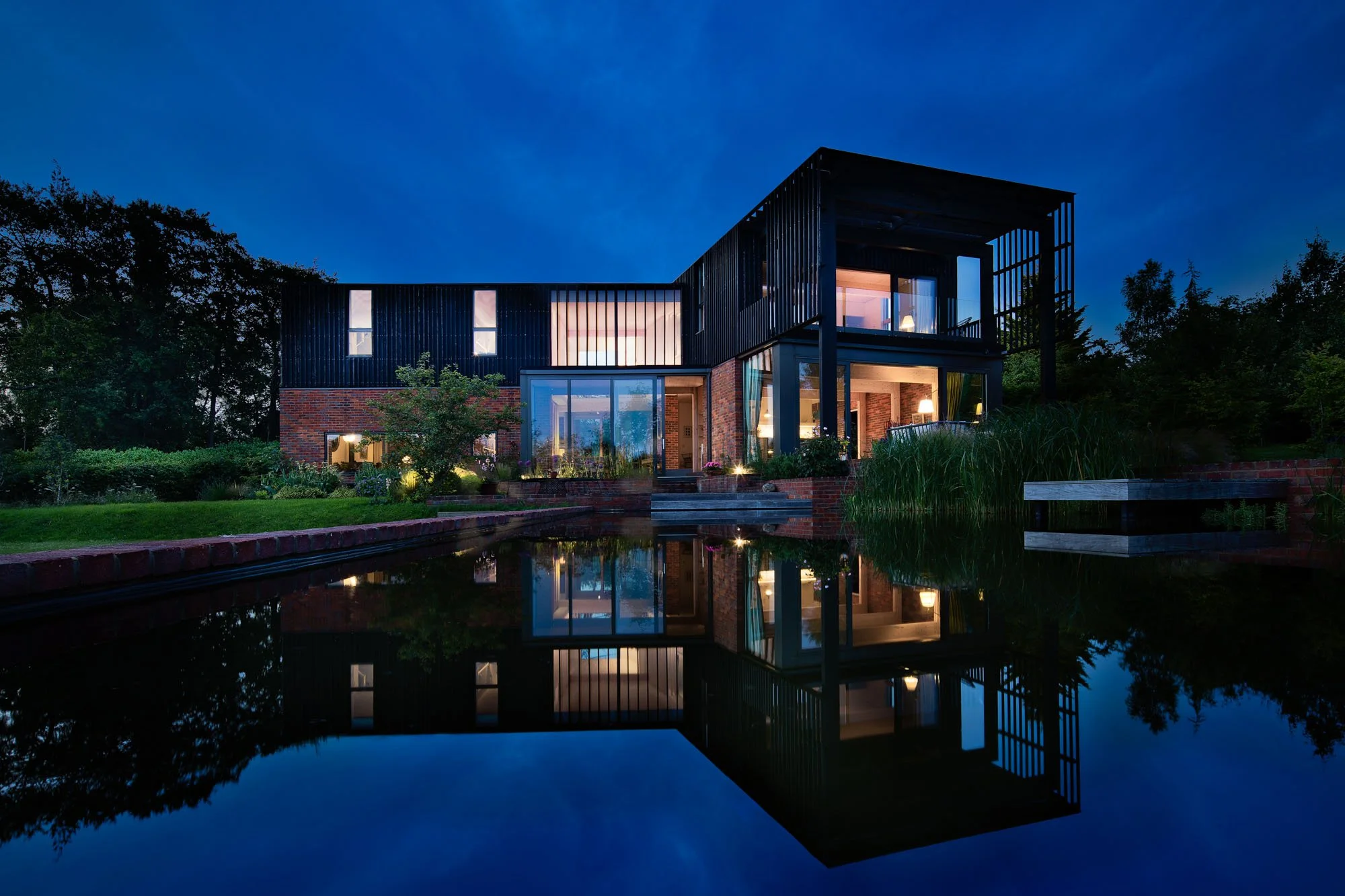 Levitate Architects — Dusk view of the exterior of Levitate's mid-century modern inspired two-story house with black timber and brick cladding, with a natural swimming lake