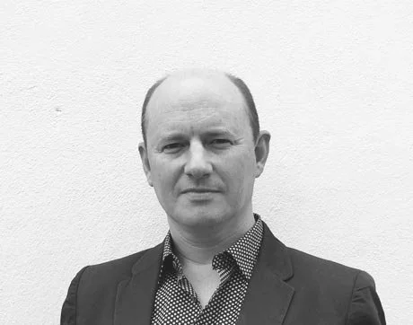 Black and white portrait of a man wearing a suit and a patterned shirt, standing against a plain white background.