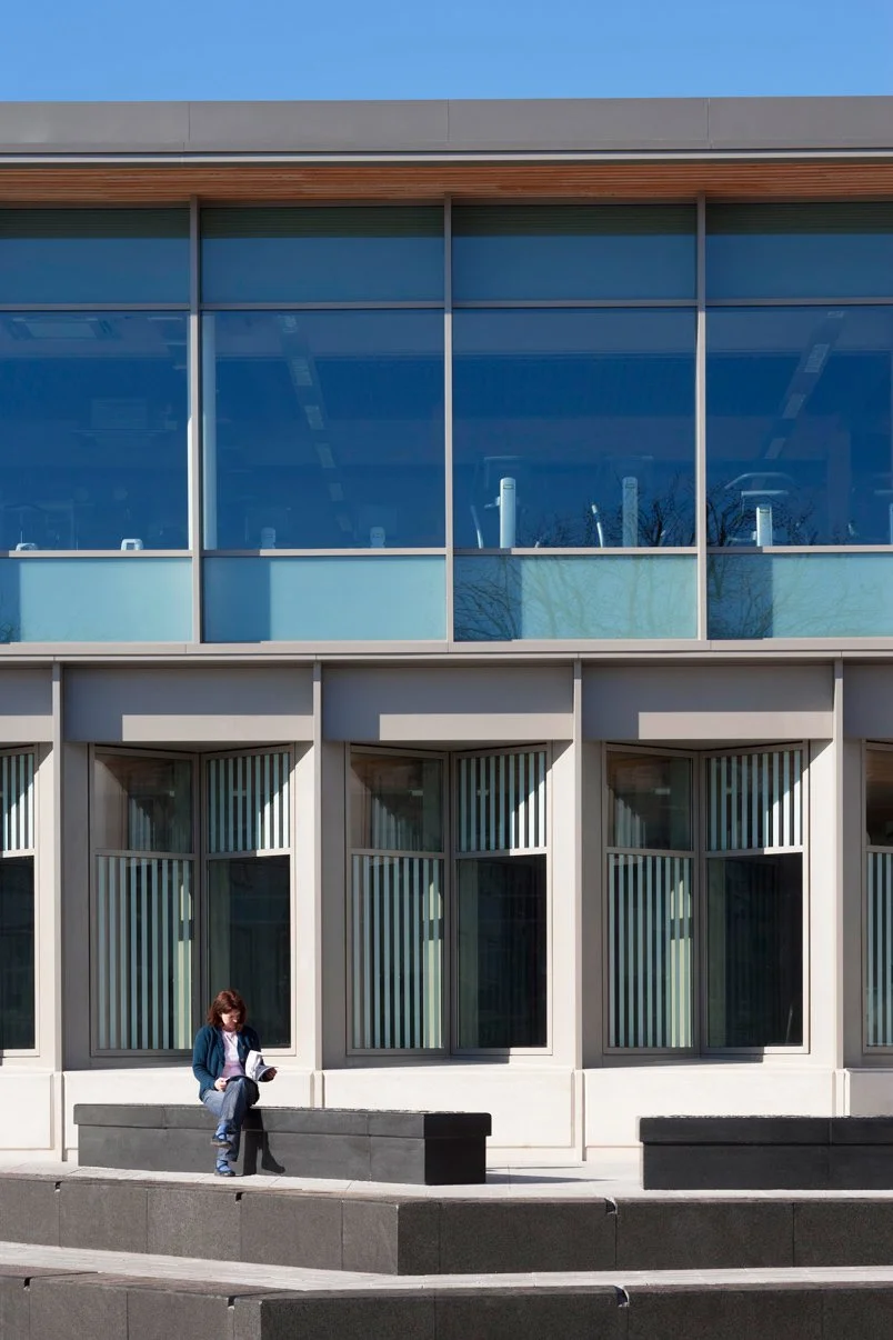 Levitate Architects' Victoria Leisure Centre: A close-up view of the contemporary facade, showing a person sitting and reading on a low stone wall outside the double-height glass windows of the gym and fitness studio.