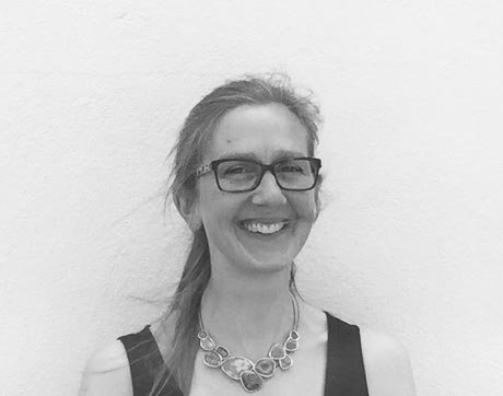 Black and white photo of a smiling woman with glasses, wearing a necklace and sleeveless top, standing against a plain wall.