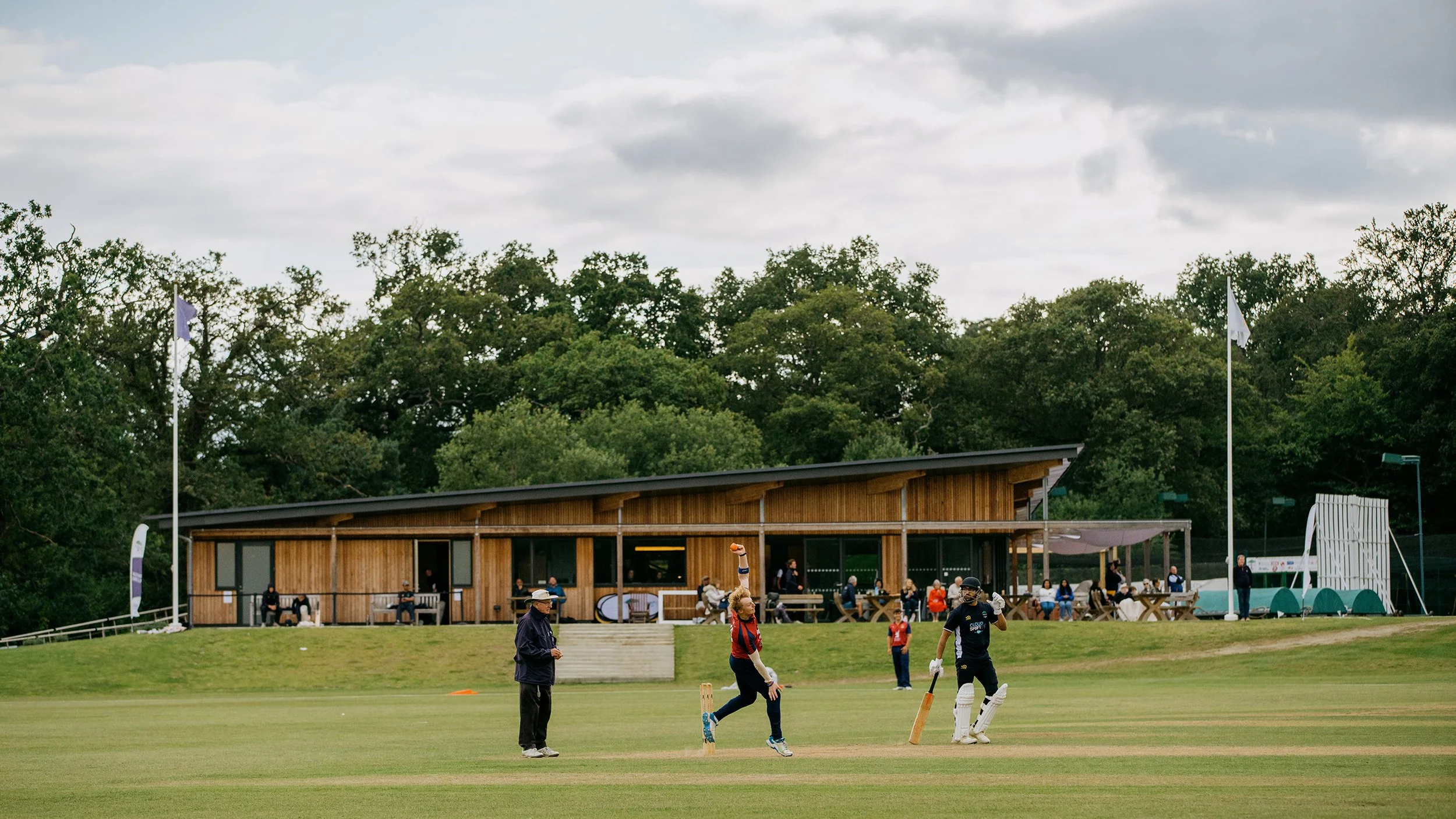 Levitate Architects' Valley End: An exterior shot of the contemporary pavilion clubroom building looking up towards the dark timber-clad upper story and roof layout from the open grounds.