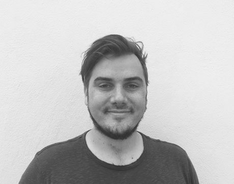 Black and white photo of a young man with short, dark hair, a light beard, and a slight smile, standing in front of a plain wall.