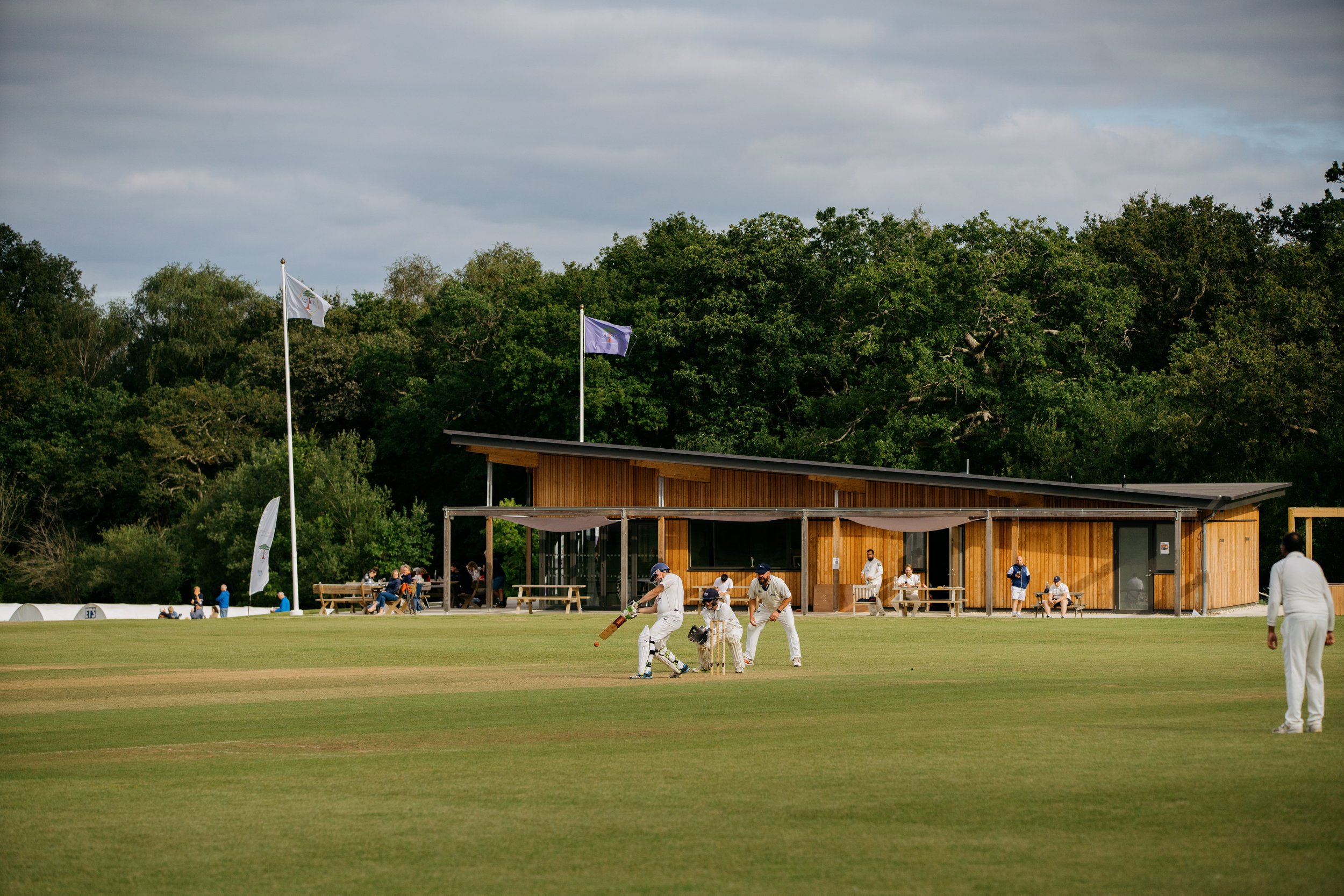 Levitate Architects' Valley End cricket pavilion: A wide view of the contemporary cricket pavilion featuring timber cladding, large glass doors, and a stone patio area facing the grounds.