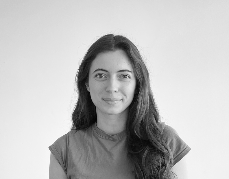 A young woman with long dark hair and a slight smile, wearing a casual T-shirt, standing against a plain wall.