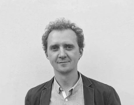 Black and white portrait of a man with curly hair wearing a blazer and collared shirt in front of a plain wall.