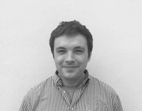 Black and white photo of a young man with short, dark hair, wearing a checkered shirt and smiling at the camera, standing against a plain white wall.