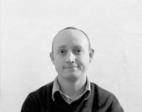 A black-and-white photo of a David, wearing a collared shirt and dark sweater, smiling slightly, standing against a plain wall.
