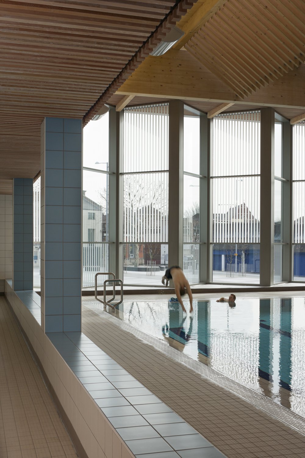 Levitate Architects' Victoria Leisure Centre: A swimmer mid-dive into the indoor pool, framed by blue tiled columns and a double-height window facade with vertical privacy louvres.