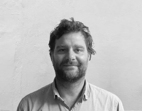 Black and white photo of a man with a beard and curly hair, wearing a collared shirt, standing against a plain wall.