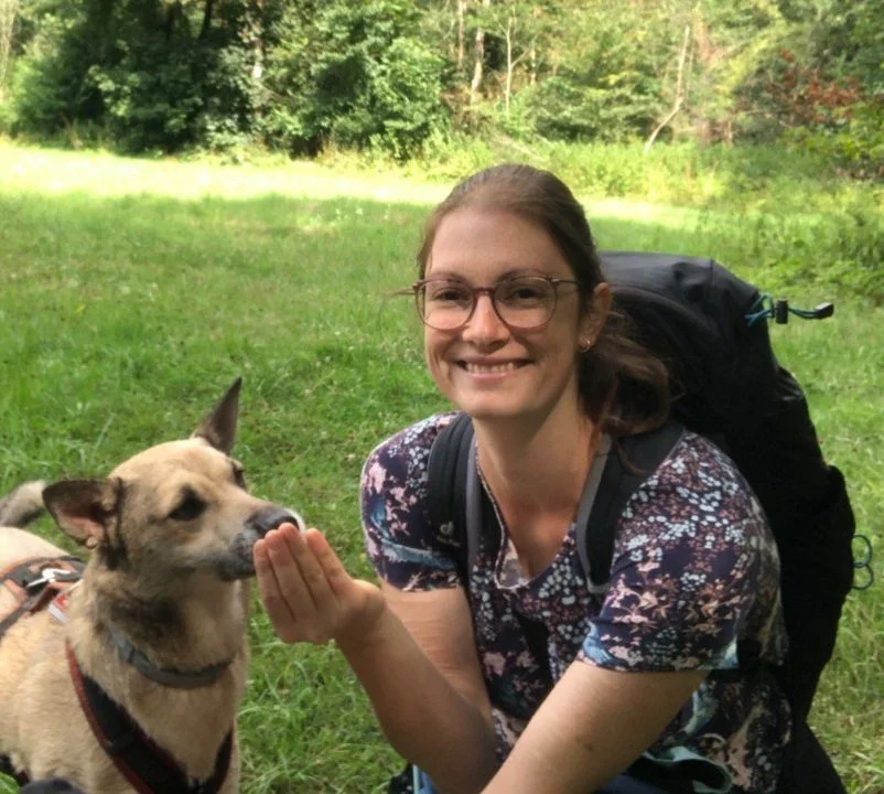 Rebekka Fuchs, Hundetrainerin in Leipzig, sitzt lächelnd auf einer Waldwiese und gibt ihrer Hündin Lupa eine Futterbelohnung aus der Hand.
