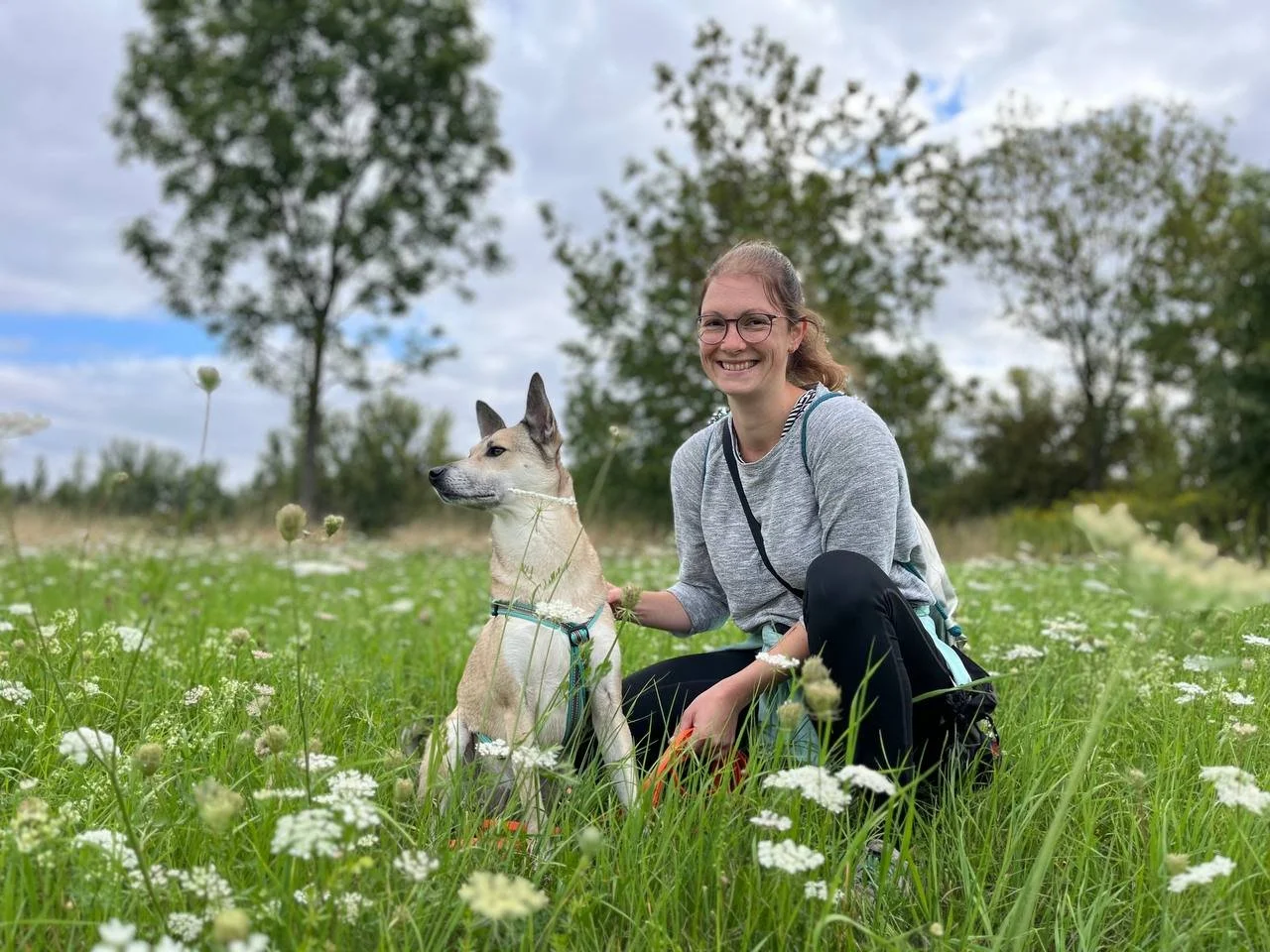Rebekka Fuchs, Hundetrainerin in Leipzig, sitzt lächelnd im Gras auf einer Blumenwiese, neben ihr ihre Hündin Lupa mit türkisem Brustgeschirr.
