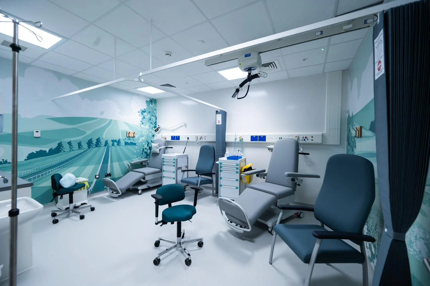 Empty hospital room with medical chairs, chairs, and equipment, with a mural of a scenic landscape on the wall.