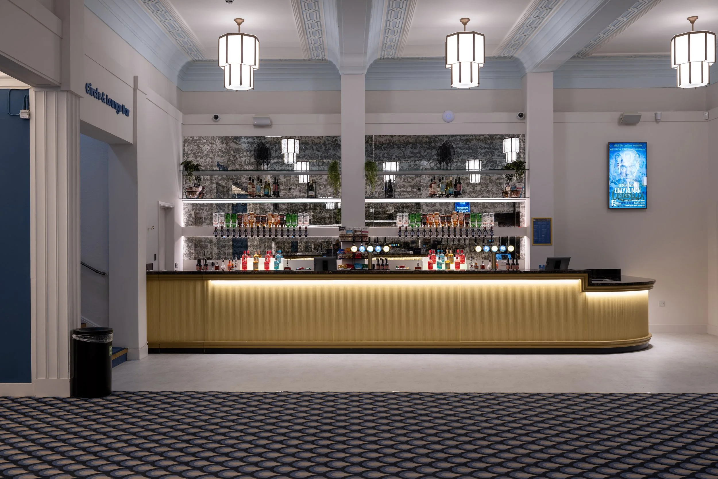 A modern hotel bar with a curved counter, bottles of alcohol displayed on illuminated shelves, and hanging pendant lights on a high ceiling with decorative molding.