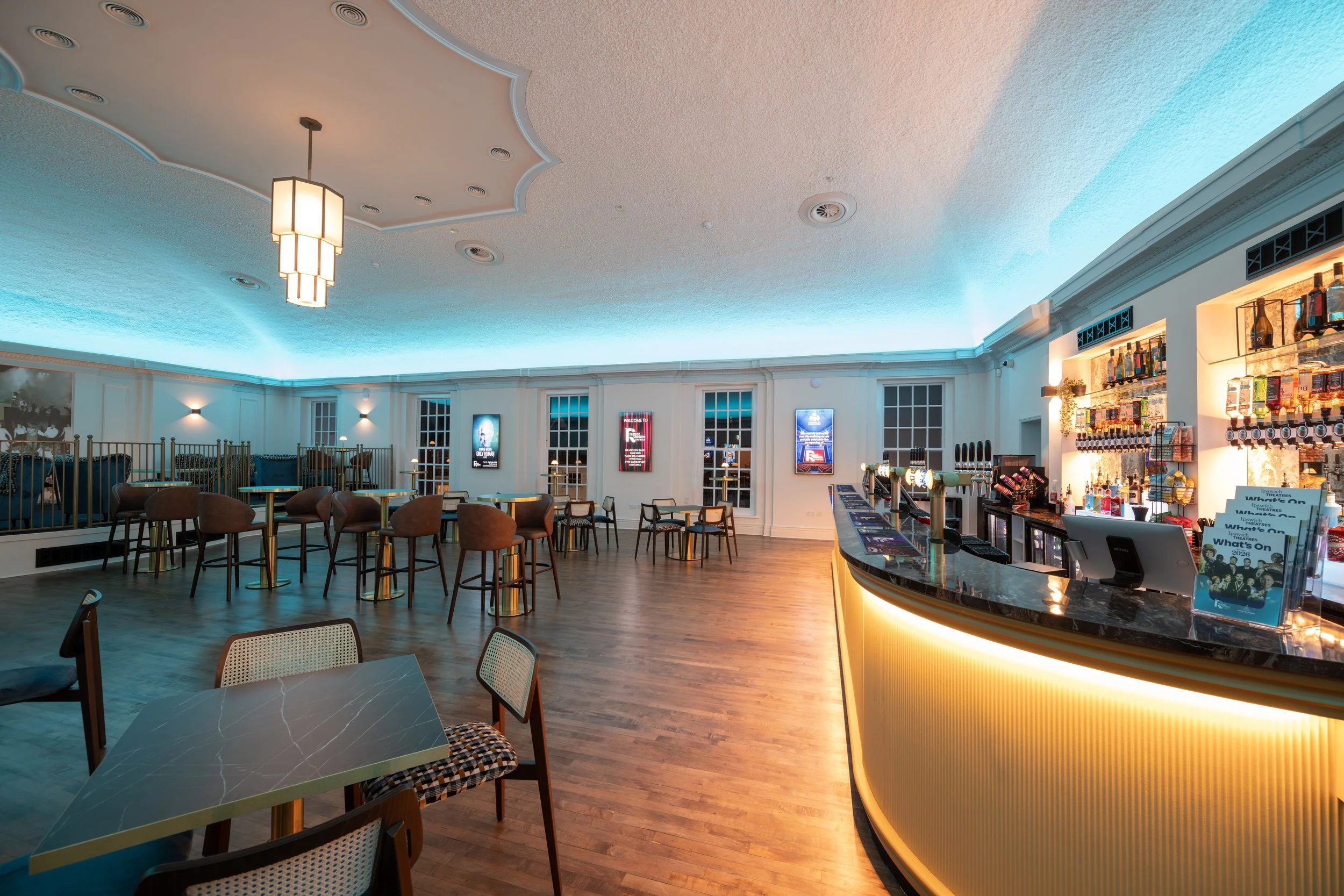 Interior of a modern bar and lounge area with a curved bar counter illuminated with warm lighting, surrounded by contemporary chairs and tables. The background features a white wall with large windows, digital screens, and shelves stocked with beverages. The ceiling has an artistic design with embedded lighting and a large pendant light.