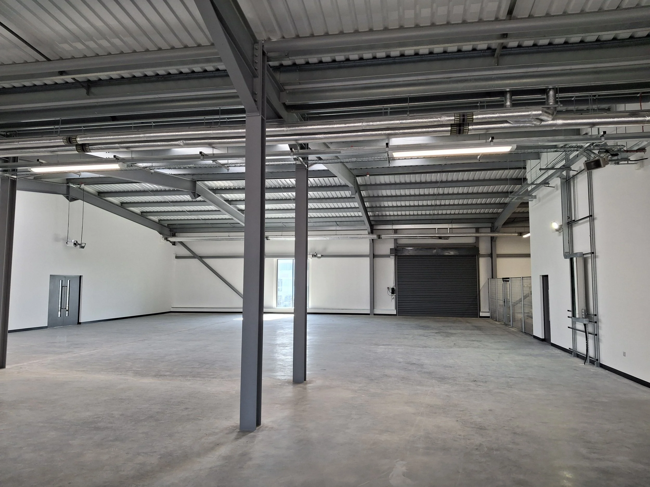 Empty industrial warehouse interior with metal pipes and beams, concrete floor, white walls, and large garage door.