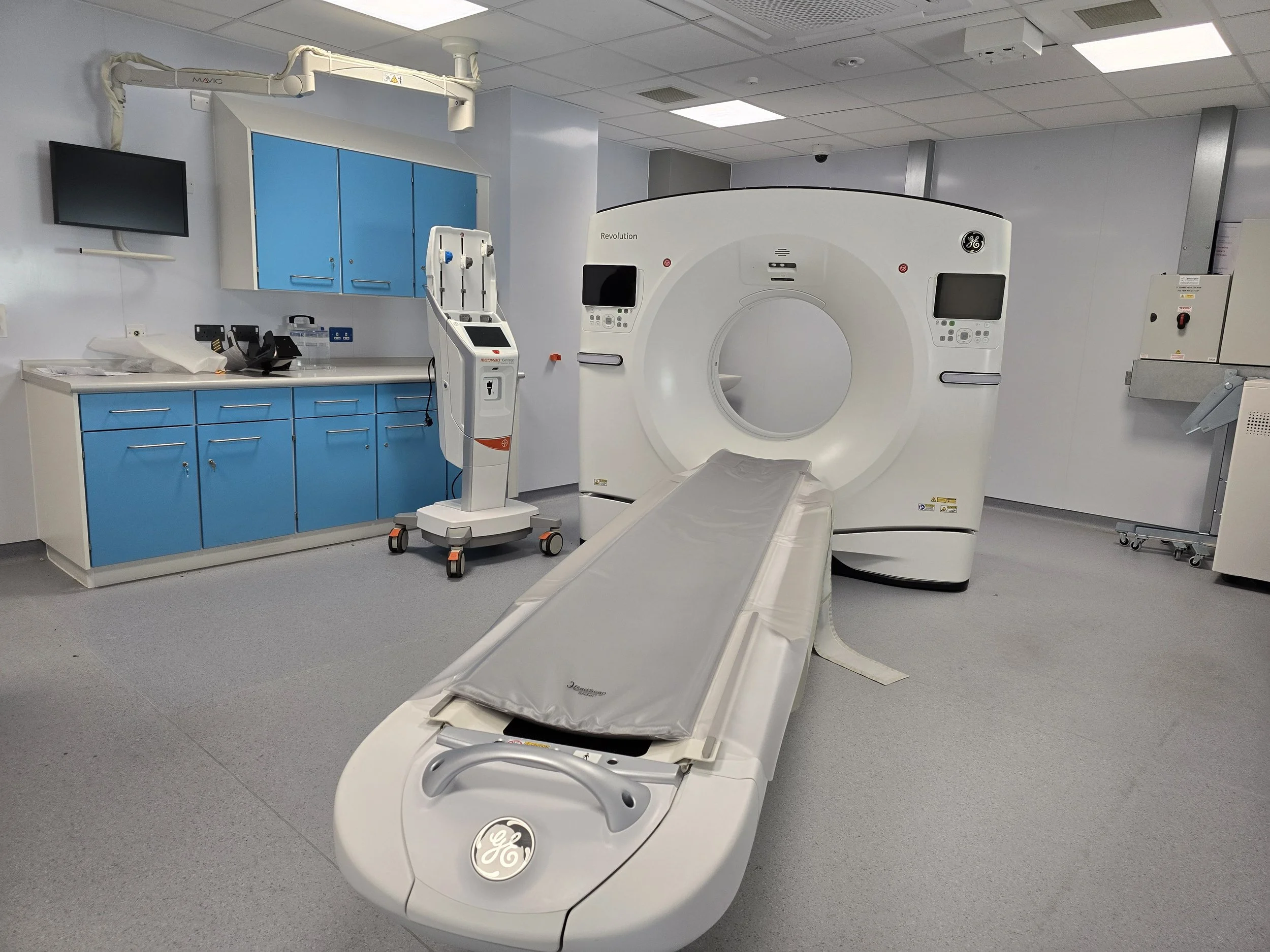 Medical imaging room with a large GE Revolution CT scanner, a patient table, blue cabinets, a monitor, and medical equipment.