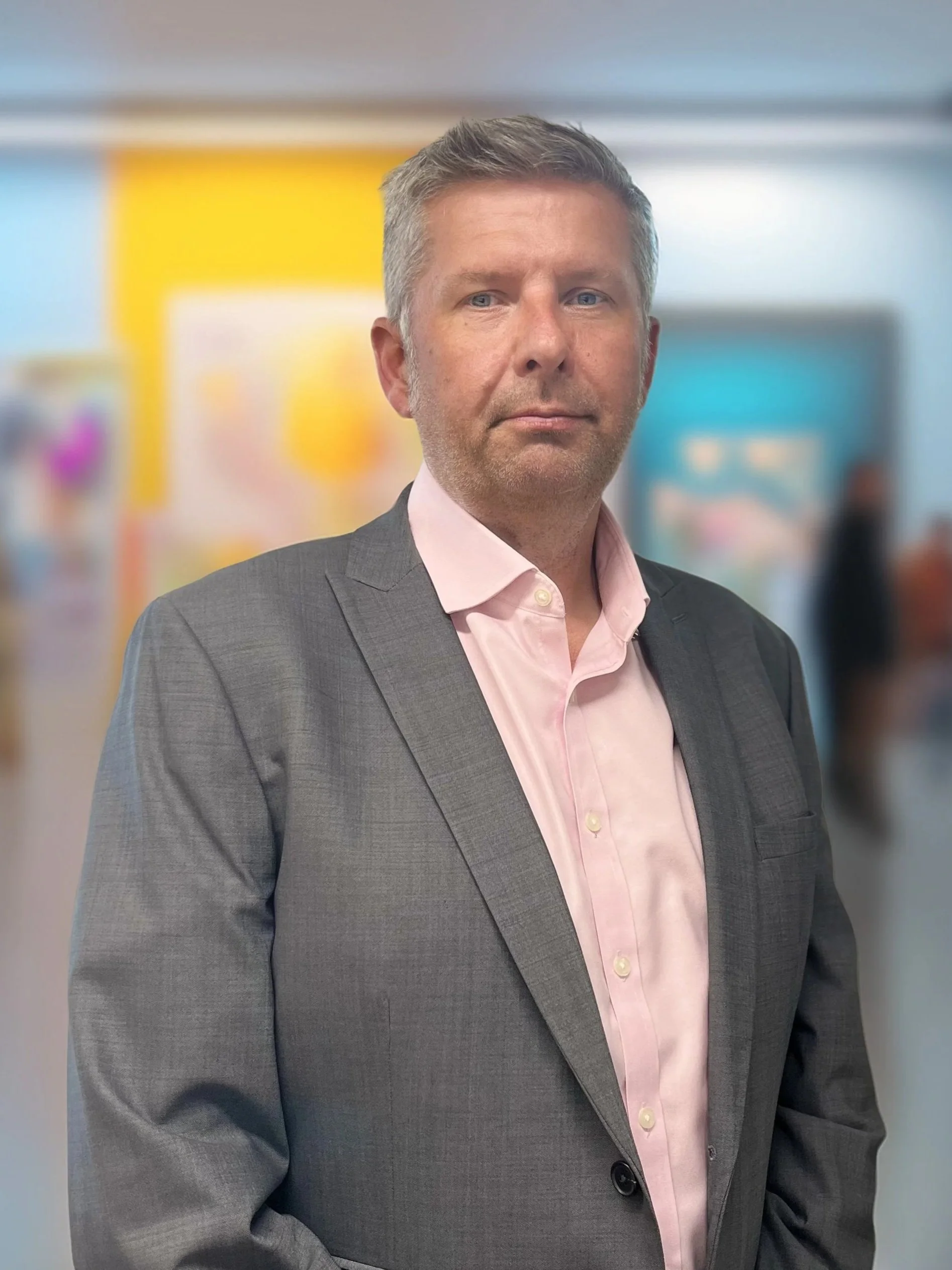 A professional man in a gray suit and pink shirt, standing in an office environment with colorful blurred backgrounds.
