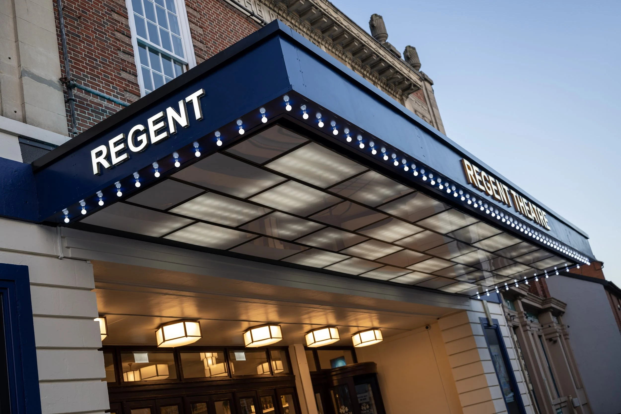 The marquee of Regent Theatre with a blue awning and white illuminated letters spelling 'REGENT'. The theatre's entrance is illuminated with warm lights and the building features brick and stone architecture.