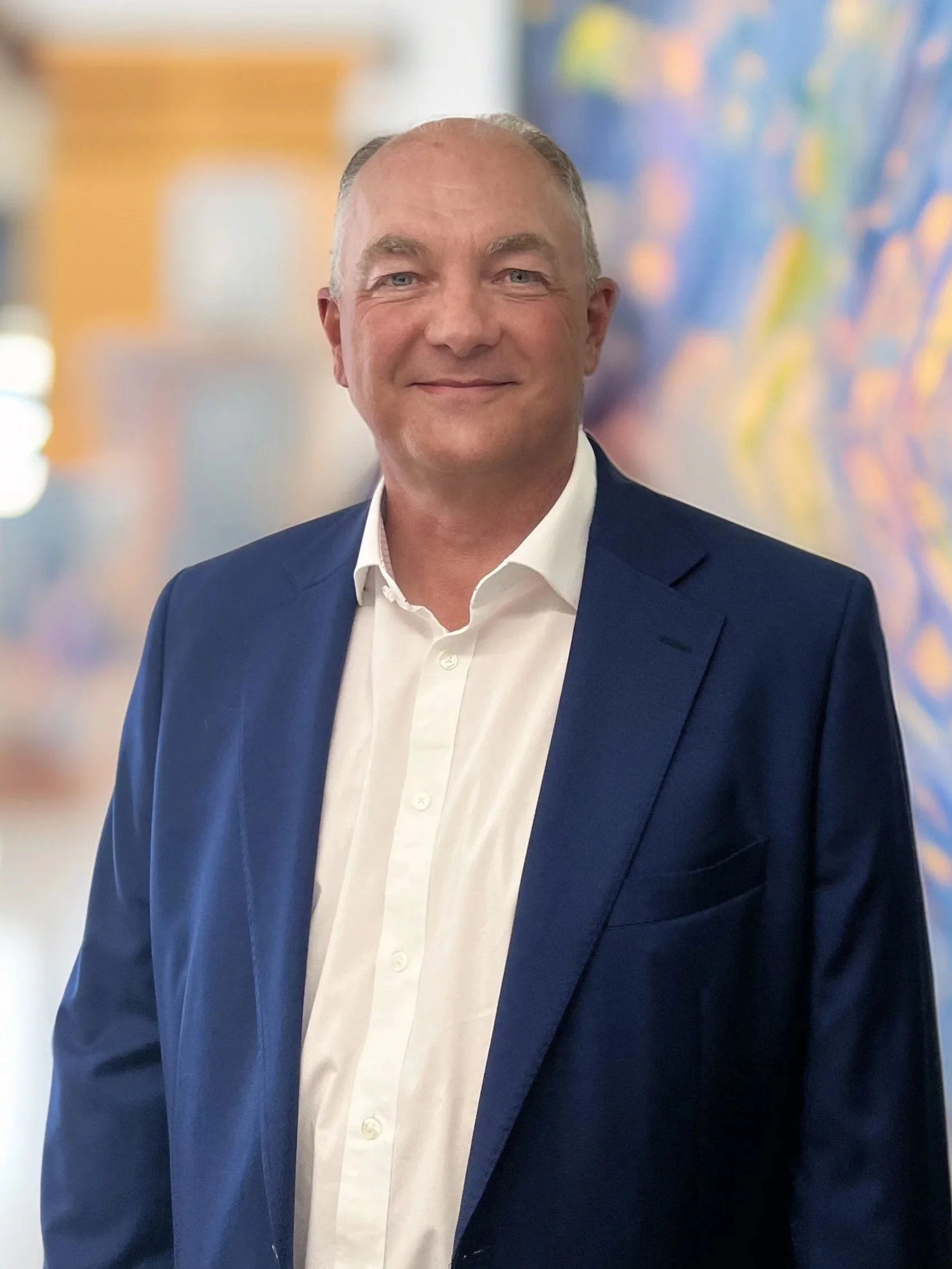 Portrait of a middle-aged man with short gray hair, wearing a navy blue suit and white shirt, smiling in an indoor setting with blurred colorful artwork in the background.