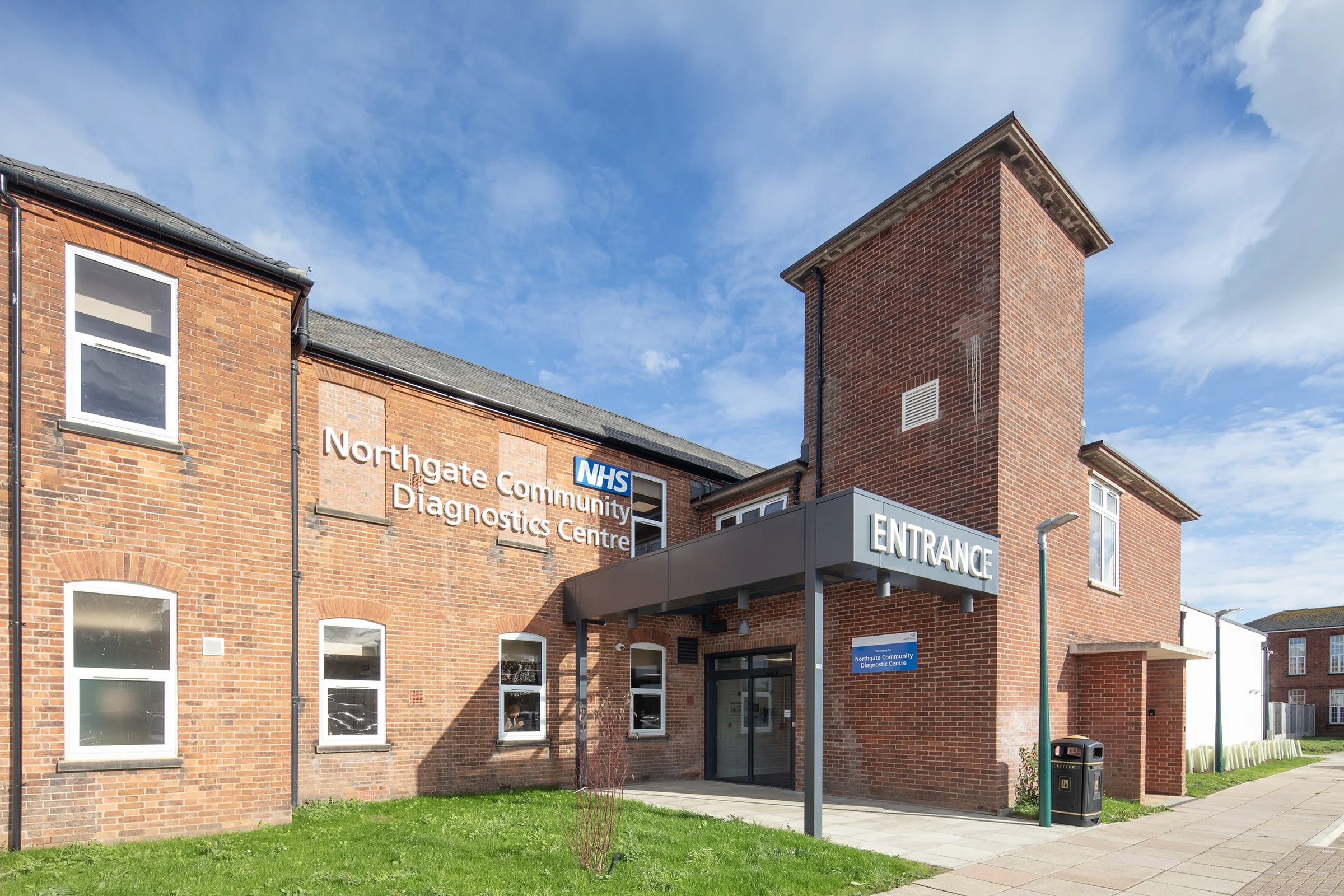 Exterior of the Northgate Community Diagnostic Centre building with brick walls, multiple windows, a sign indicating the entrance, and a clear blue sky.