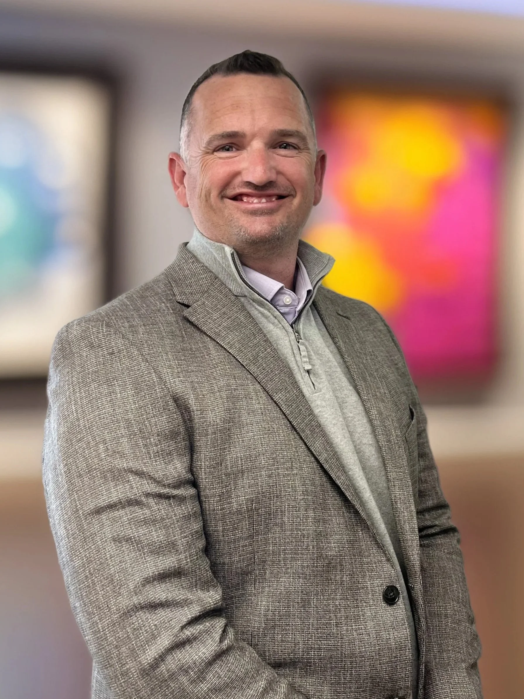A man smiling and wearing a grey blazer over a zip-up shirt, standing in front of colorful, blurred background art.