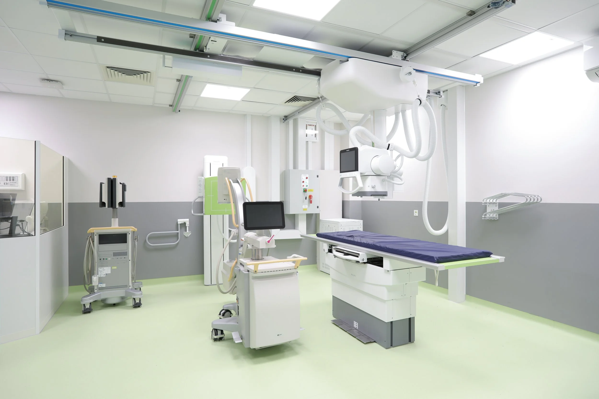 A hospital room with medical equipment including an examination bed, monitors, and ventilator apparatus on a light green floor and white walls.