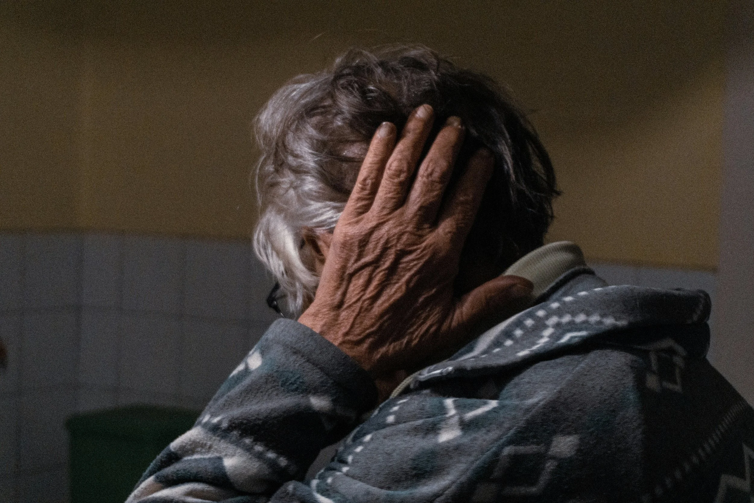 Old person with headaches with gray hair and a dark skin hand covering their face, wearing a dark patterned sweater.