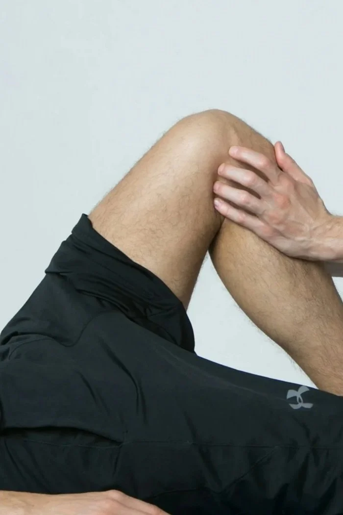 A person with one hand on their knee, sitting against a plain white wall, wearing black athletic shorts and a black athletic shirt.