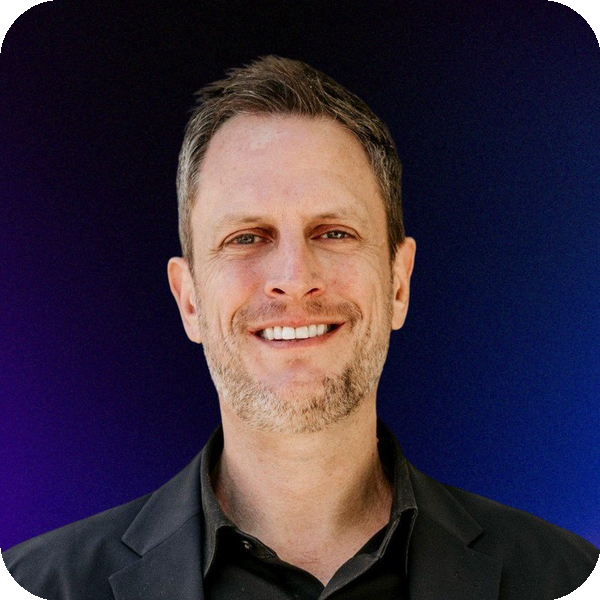 Adam Harris, A man with short brown hair and a beard, smiling, wearing a dark suit jacket and black shirt, against a blue gradient background.