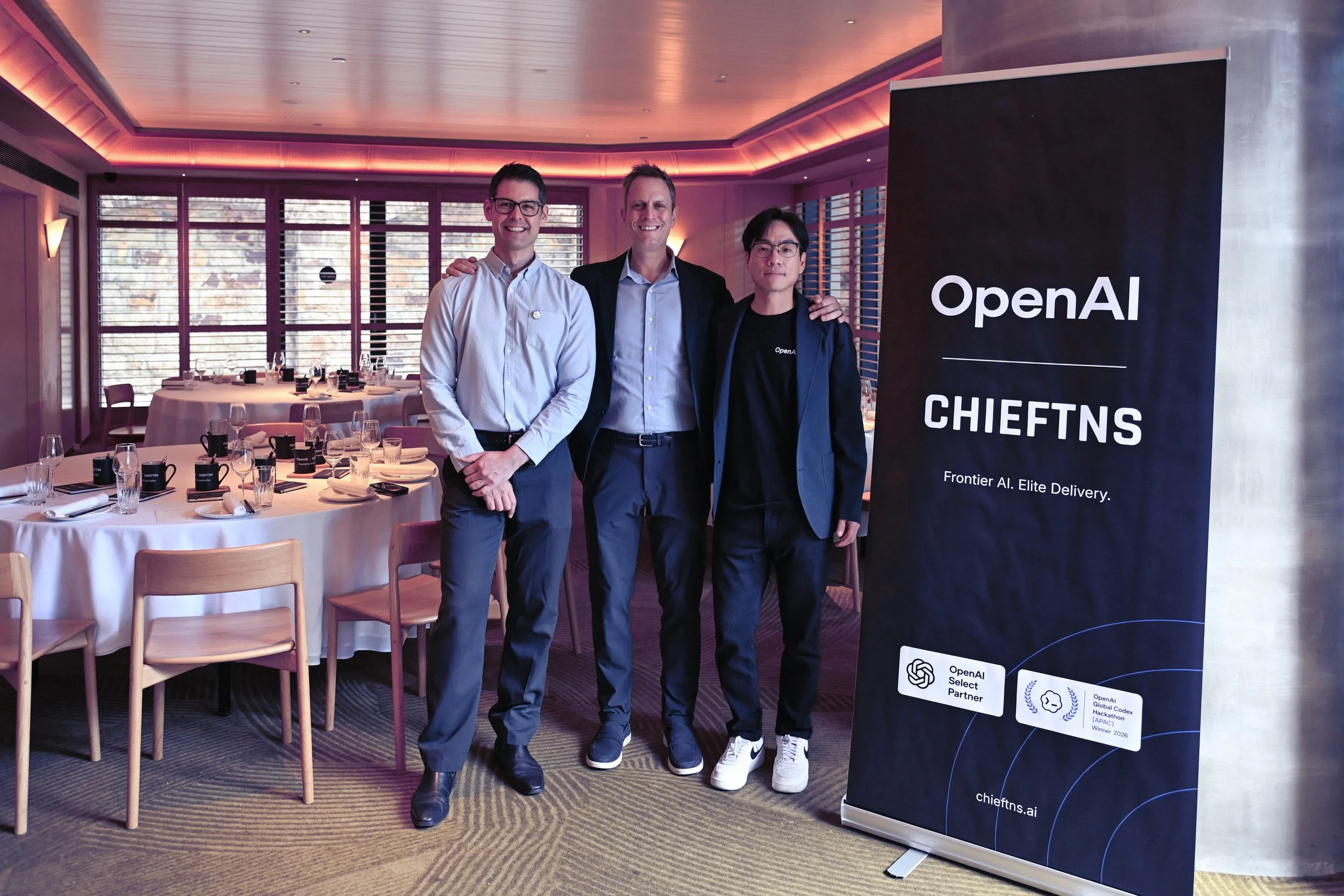 Three men standing together in a restaurant or conference room near a large vertical banner that reads 'OpenAI CHIEFTNS' and 'Frontier AI. Elite Delivery.' The men are smiling and dressed in business casual and smart casual attire. The restaurant has round tables set with glasses, plates, and utensils, and the room features large windows with shutters and purple-pink lighting.