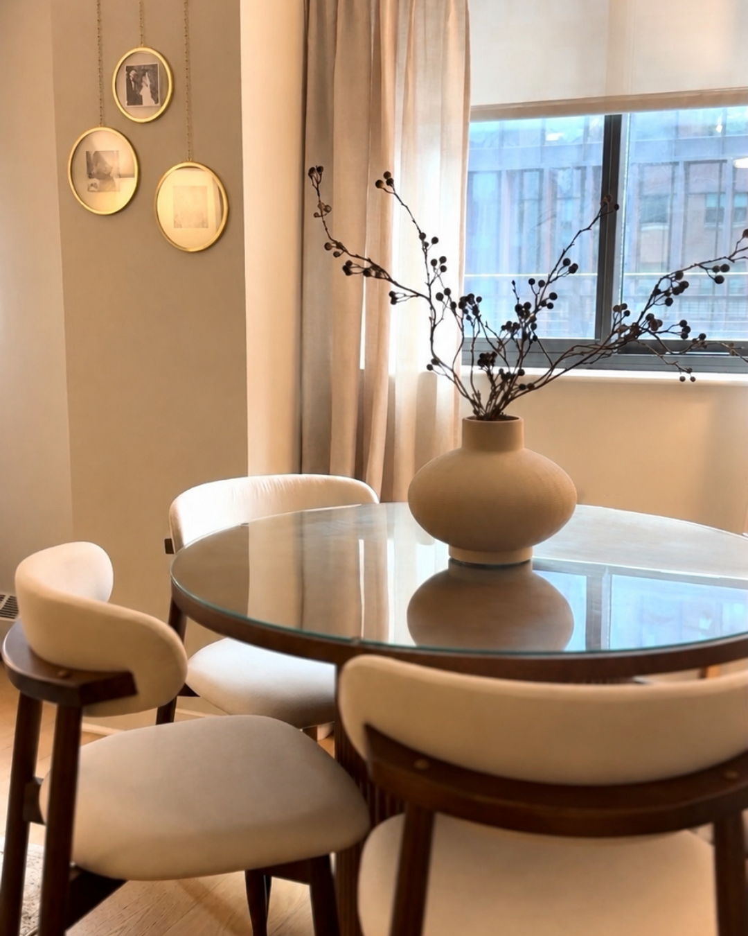 A round glass dining table with a beige vase and decorative branches, surrounded by white upholstered chairs, in front of a window with beige curtains and a wall with three gold-framed photographs.