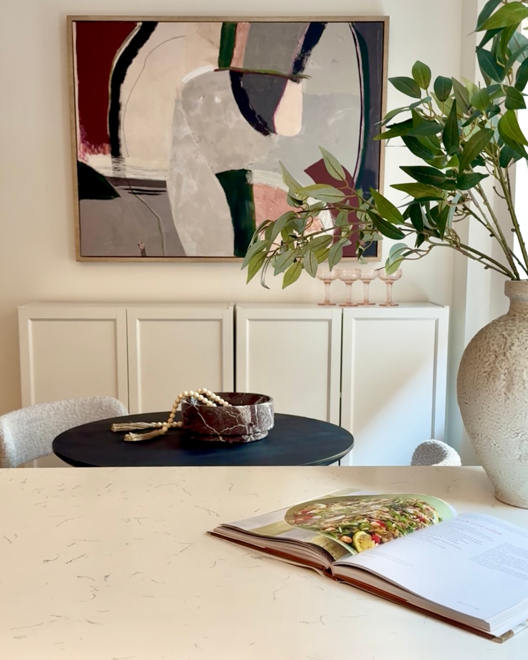 A modern dining room with a white countertop, open cookbook, large vase with green leafy plant, and abstract artwork on the wall. A small black table with a red marble bowl and pearl necklace is in the background.