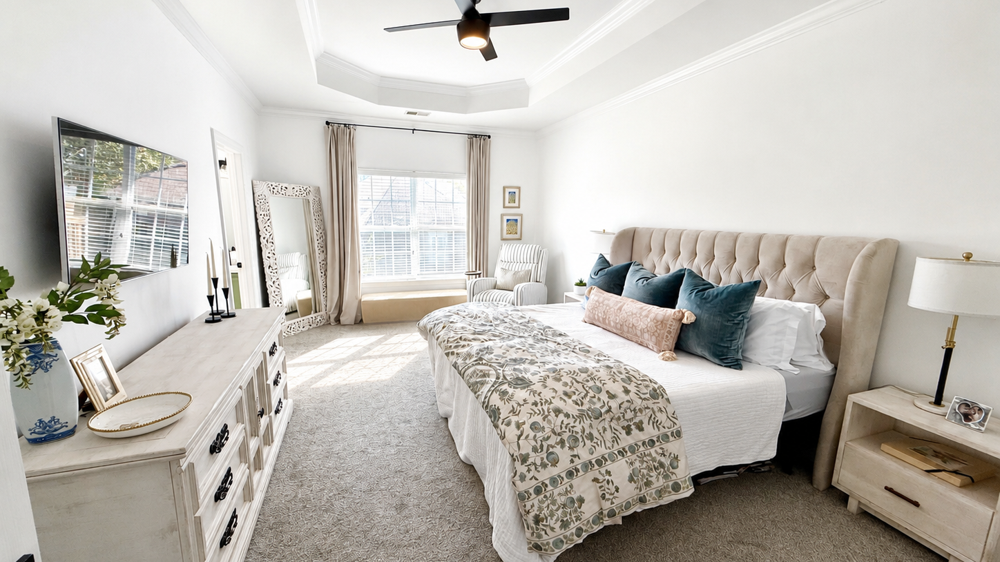 A bright, spacious bedroom with a large bed, cream tufted headboard, multiple pillows, and a patterned bedspread. Contains a white dresser, a flat-screen TV, a window with beige curtains, a striped armchair, and decorative items like a mirror and framed art.