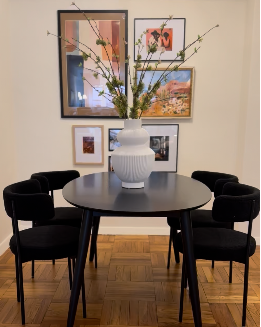 A round black dining table with six black chairs, topped with a large white vase holding spring branches with green buds, in front of a wall decorated with a collage of framed artwork.