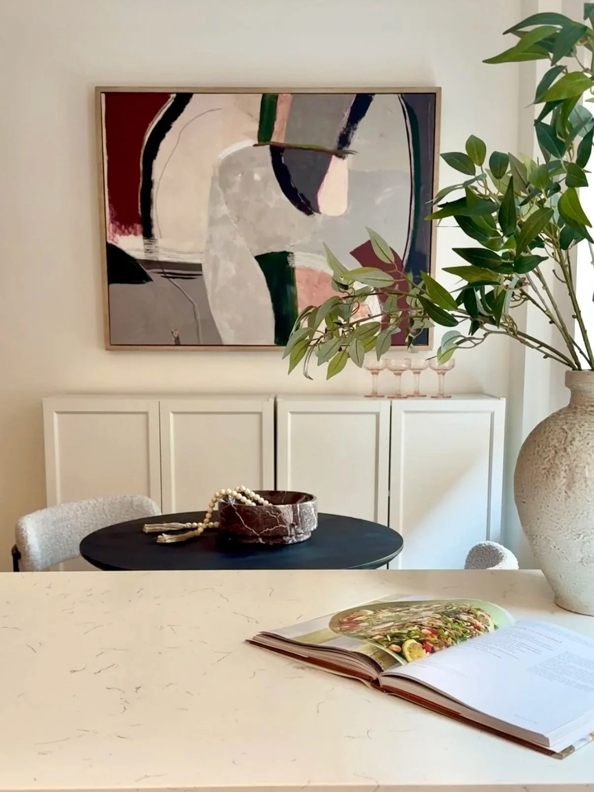 Living room with abstract wall art, a large leafy plant in a textured vase, a black round table with a marble bowl and pearl necklace, an open magazine with food, and white cabinetry.