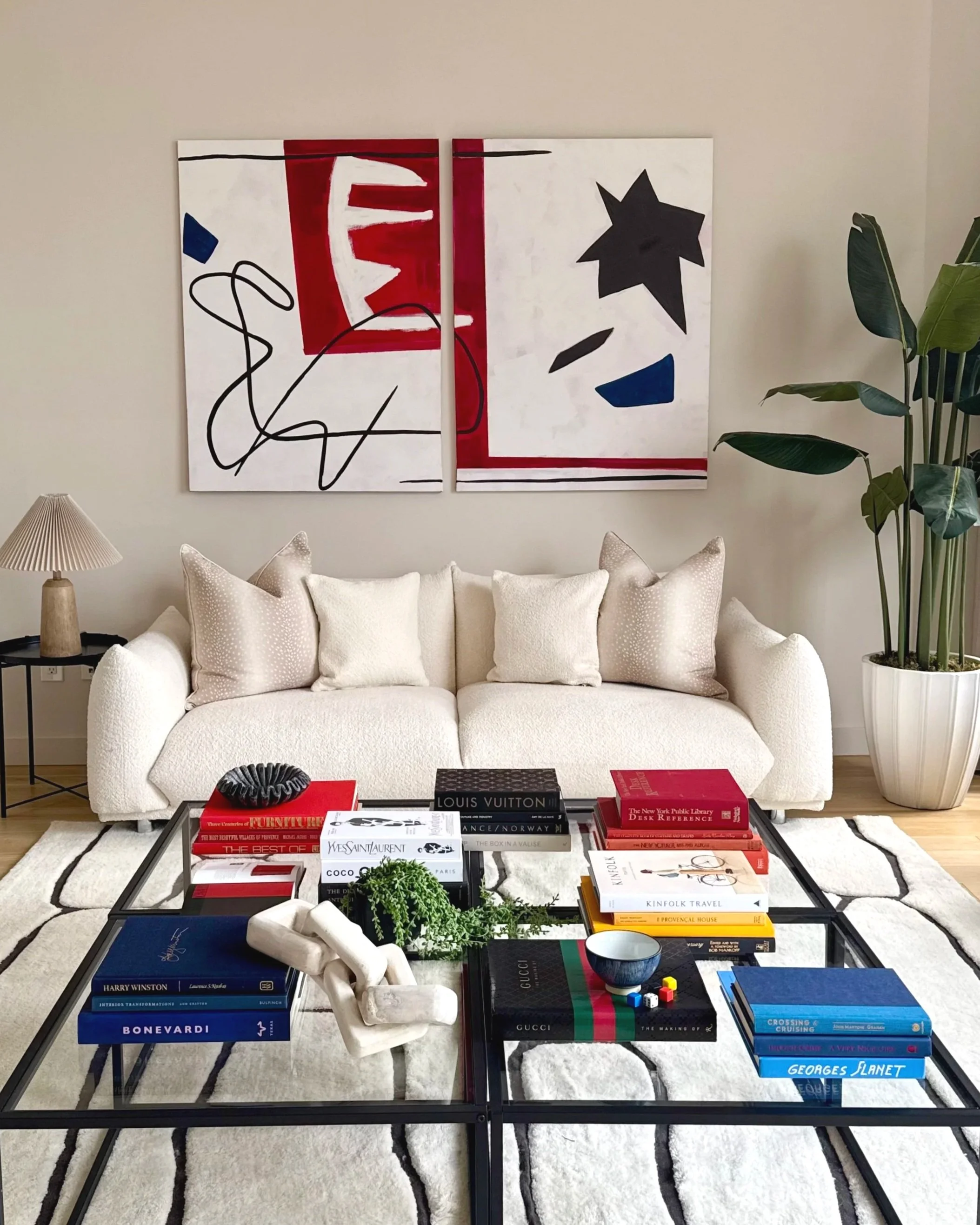 Living room with white sofa, stacks of art books on glass coffee table, abstract artwork on wall, large potted plant, beige walls, wall-mounted lamp, and decorative pillows.