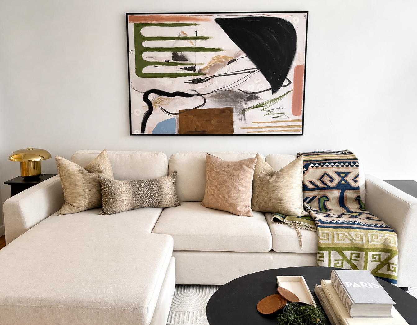 Living room with a white sectional sofa adorned with beige, taupe, and patterned pillows, a black coffee table with books and decorative items, a gold table lamp on a black side table, a colorful blanket on the right side of the sofa, a large abstrac