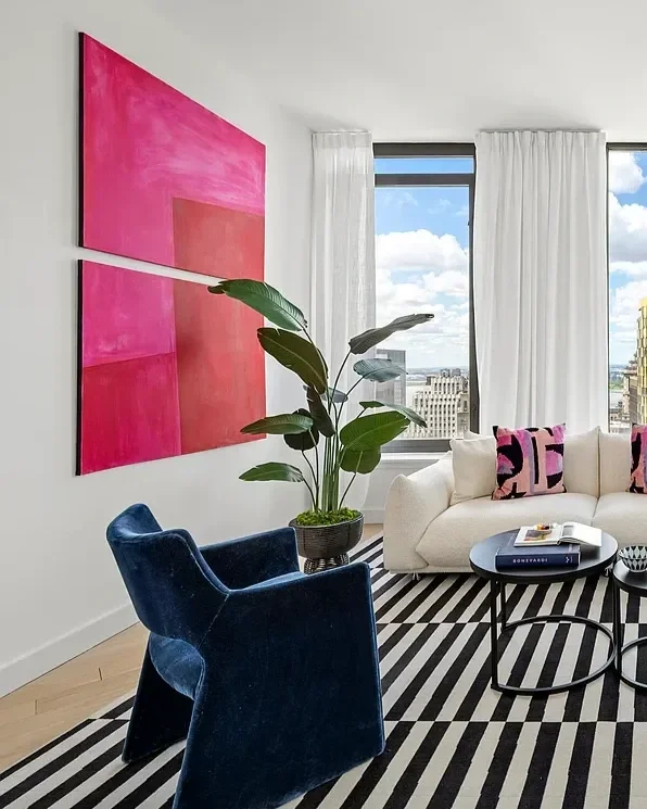 Living room with white sofa, blue velvet armchair, black round coffee table, striped black and white rug, large plant, and a pink abstract painting on the wall. Large windows with white curtains showing city buildings outside.