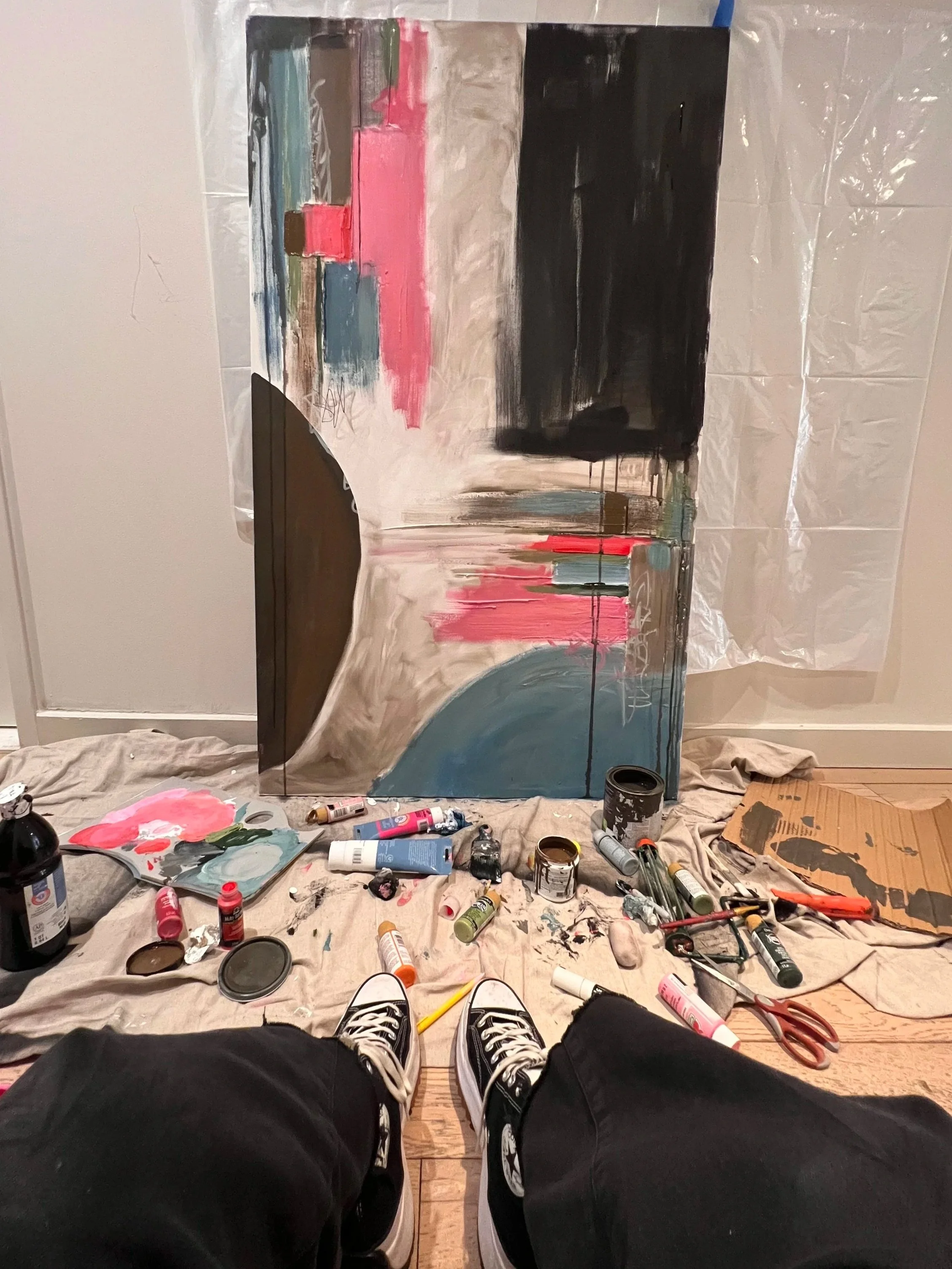 A person wearing black sneakers and black pants sitting on a wooden floor in a room undergoing painting, with an incomplete mural on the wall. The mural features abstract shapes and colors, including pink, blue, black, and beige. The table or surface
