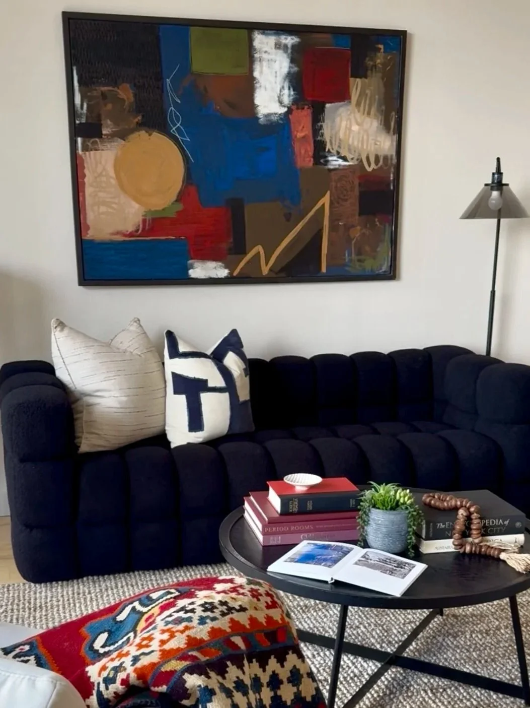 Living room with a black sofa, decorative pillows, a round coffee table with books, a small potted plant, an open photo album, and a colorful patterned blanket. An abstract painting hangs on the wall, and a floor lamp is to the right.