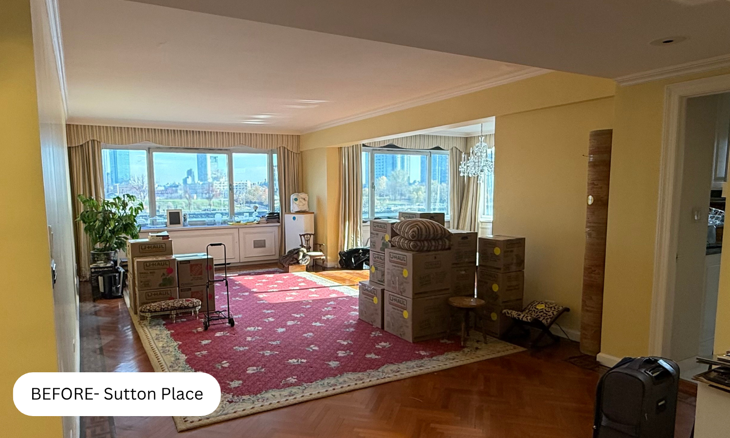 Living room filled with boxes and packed items, indicating moving preparations, with large windows showing city views in the background.