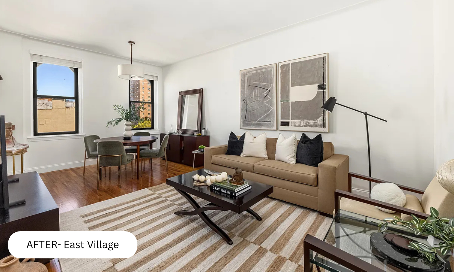 A stylish living room with a beige sofa, black and white pillows, abstract art on the wall, a black coffee table with books and decor, a round dining table with four chairs near a window, hardwood floors, a mirror, and a floor lamp.