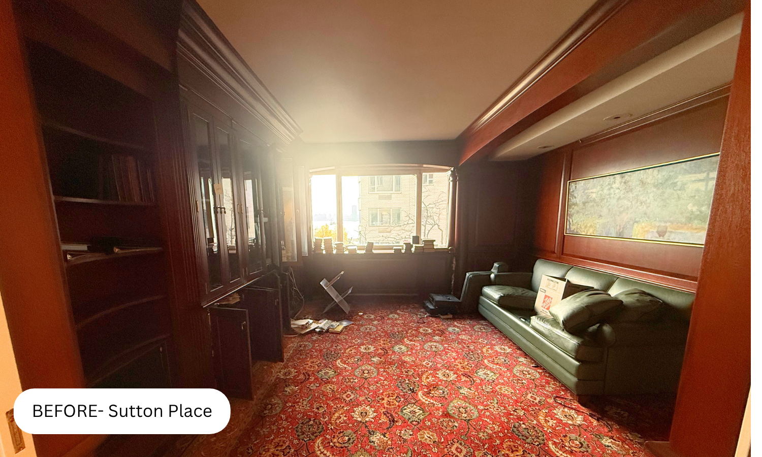 A living room with dark wood paneling, a large window, a green leather sofa with pillows, a red patterned carpet, and built-in bookshelves along one wall, with sunlight coming through the window.