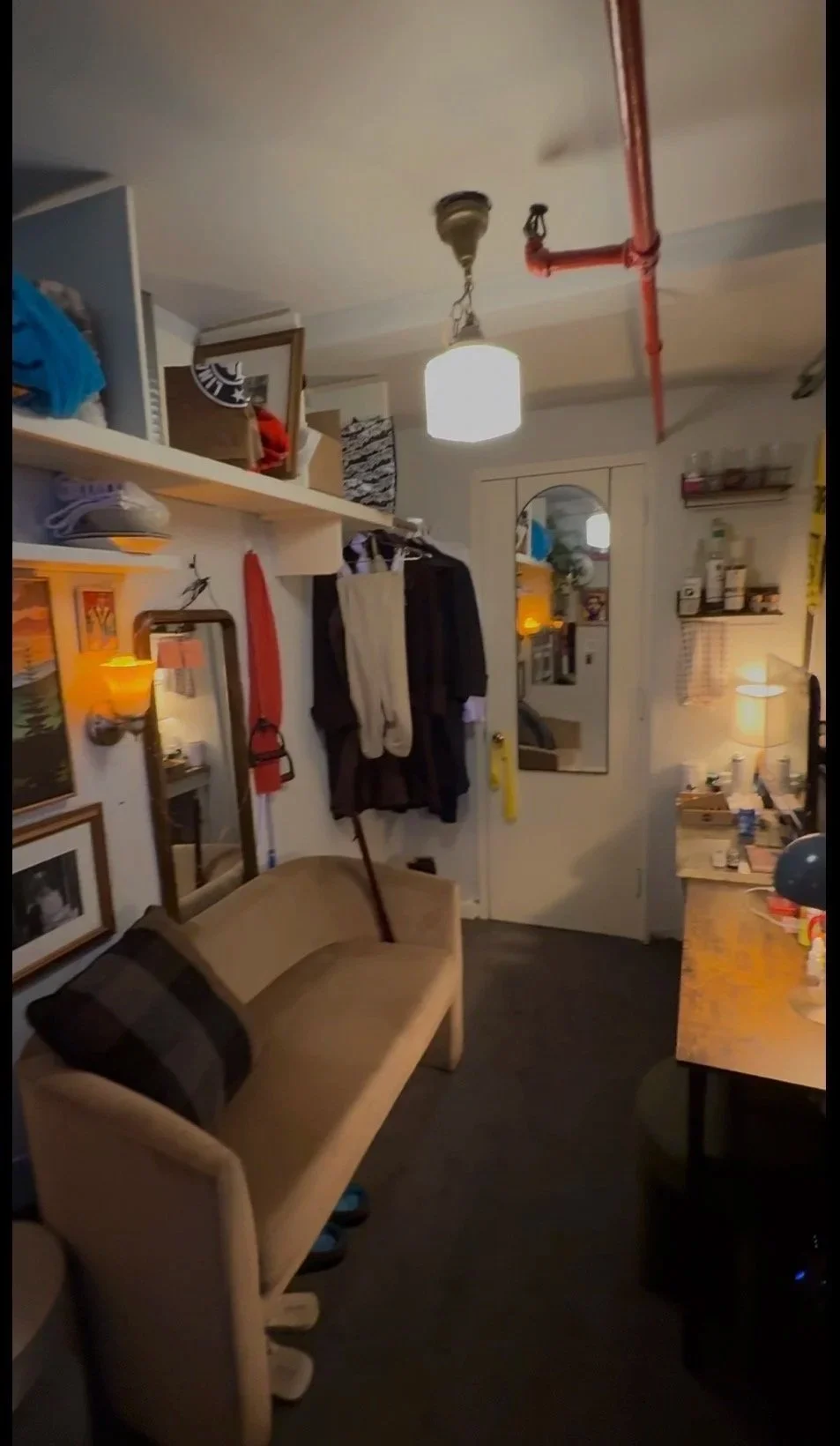 Cozy apartment room with a beige sofa, mirror, hanging clothes, a desk with a lamp, and storage shelves.