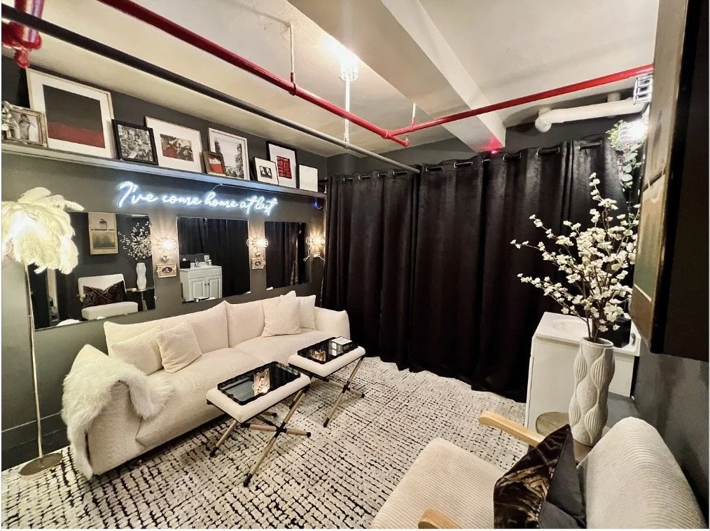 A stylish living room with a white sofa, mirrors, framed pictures on the wall, black curtains, a vase with white flowers, and a neon sign that reads 'You are home at last'.