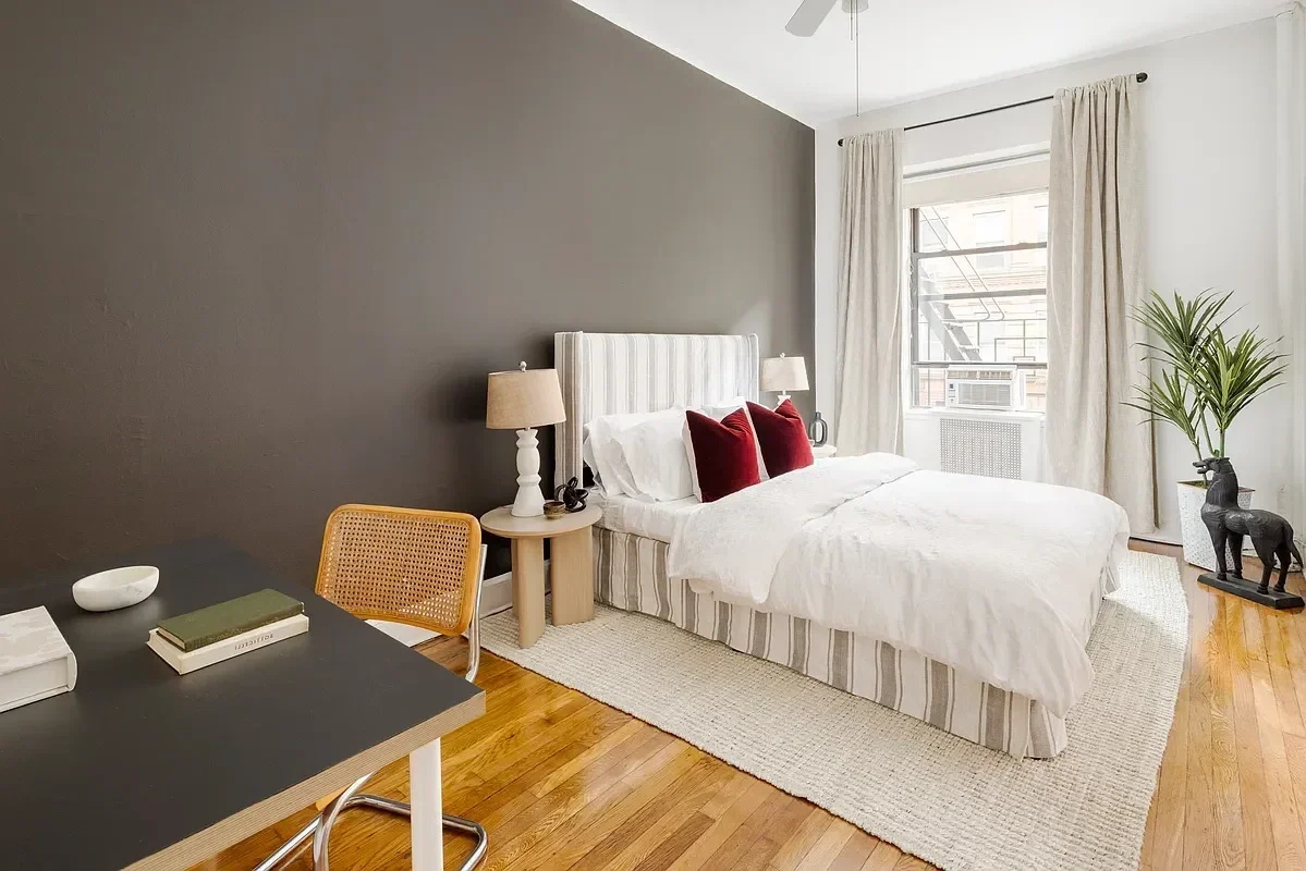 A bedroom with a white bed, two red pillows, beige nightstand with lamp, beige curtains, window with fire escape outside, potted plant, and wooden floor.