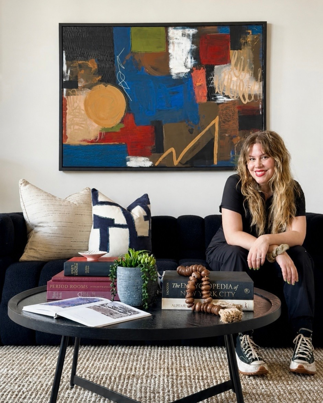 A woman with long wavy hair, sitting on a dark sofa, smiling, in a living room with a large colorful abstract painting on the wall behind her. There are decorative pillows, a table with books, a potted plant, and a beaded necklace on the table.