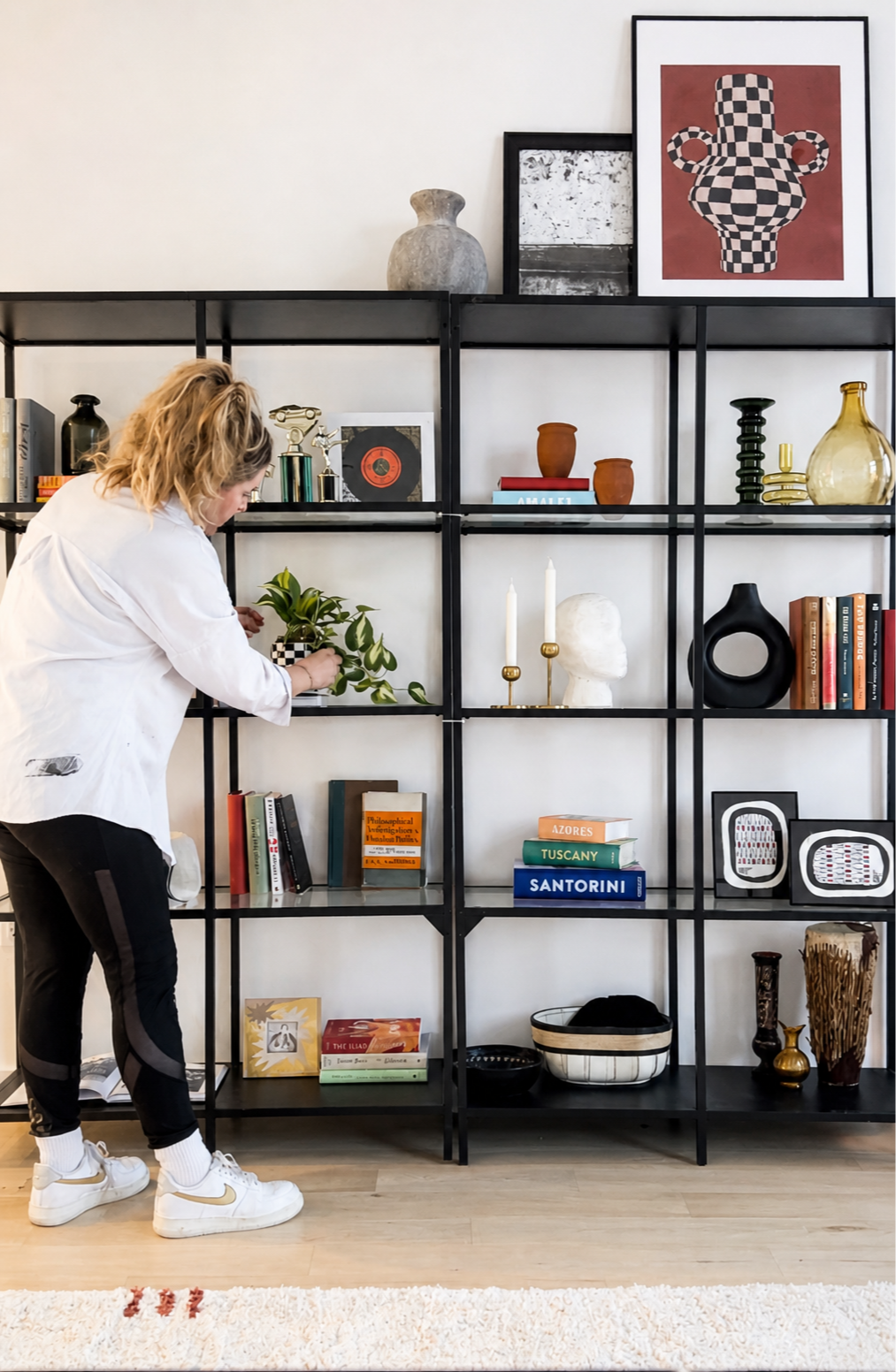 A woman with blonde hair wearing a white shirt and black pants arranging a green plant in a black and white checkered planter on a black metal shelf. The shelf contains decorative items, books, and framed artwork against a white wall.